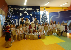 Dzieci trzymające prezenty, Pani dyrektor Maria Królikowska, pani Arleta Kalinowska i pani Ewa Janicka ustawione wokół Mikołaja, w tle dekoracja świąteczna.